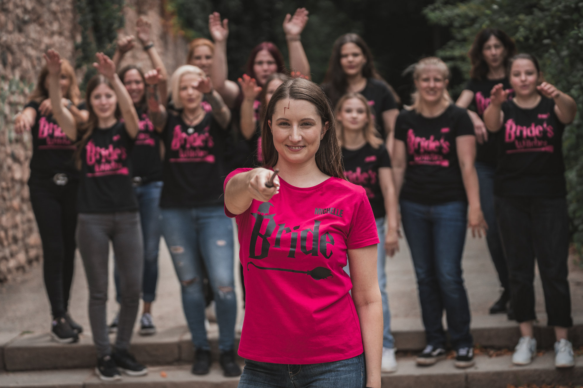 Braut im pinken Shirt zeigt in die Kamera, ihre Freundinnen jubeln dahinter