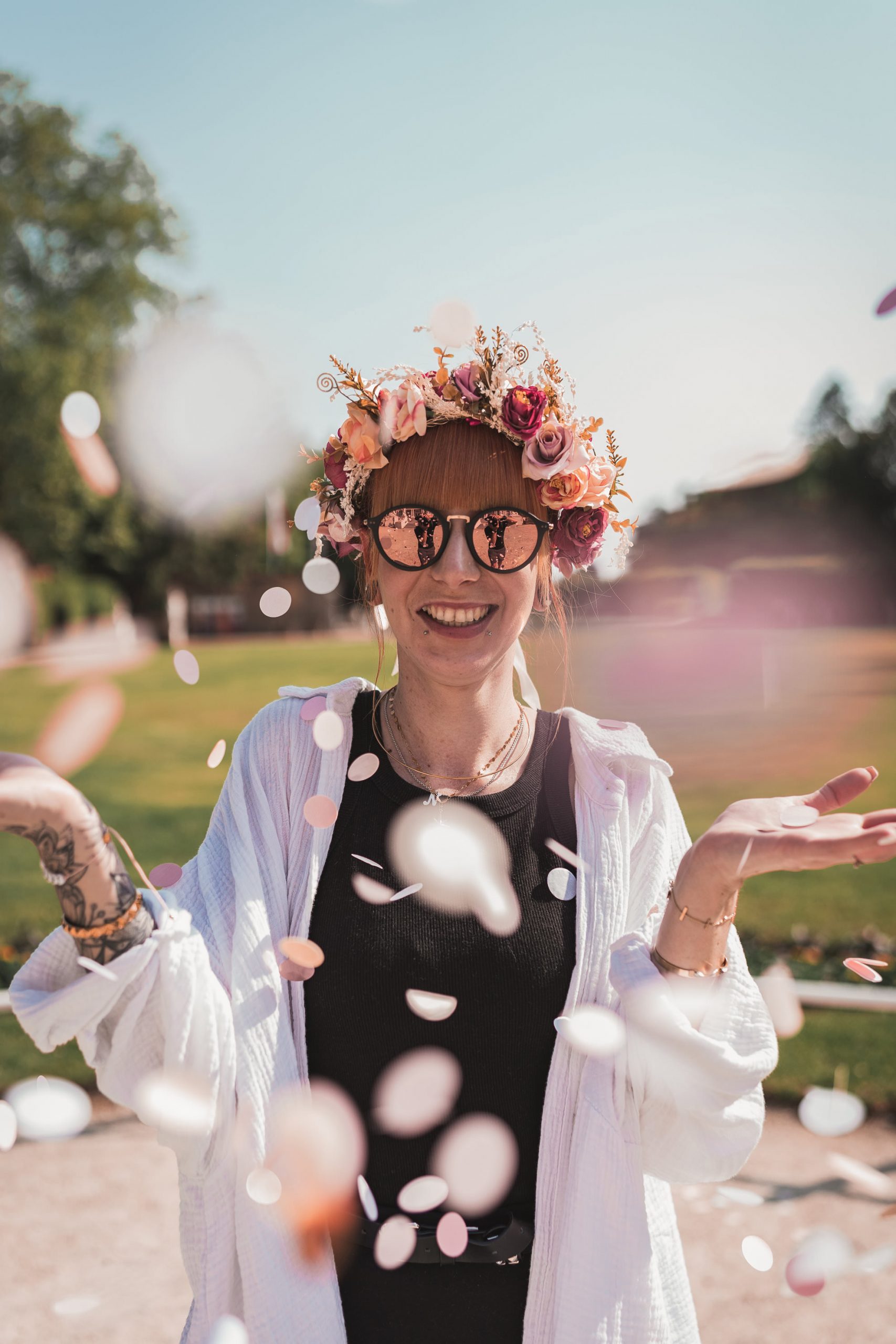 Braut mit Blumenkranz und Sonnenbrille lacht, Konfetti fliegt um sie herum
