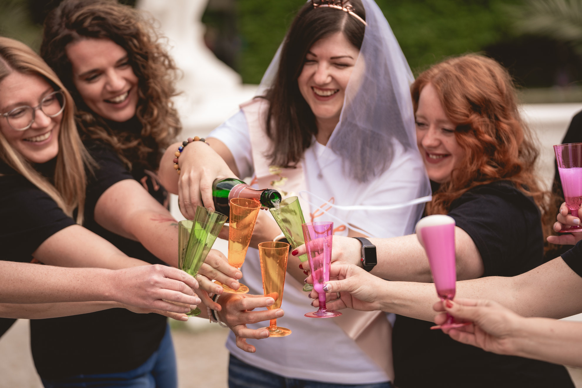Braut mit Schleier schenkt ihren Freundinnen Sekt in bunte Becher