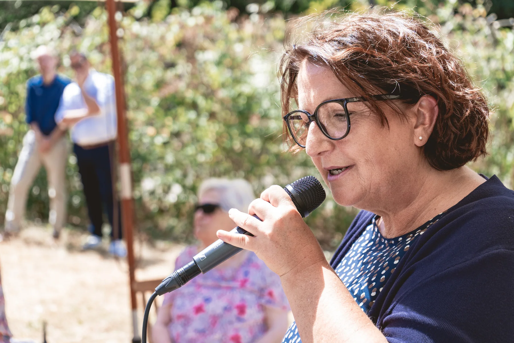 Petra Melzer spricht konzentriert ins Mikrofon, Gäste sitzen unscharf im Hintergrund