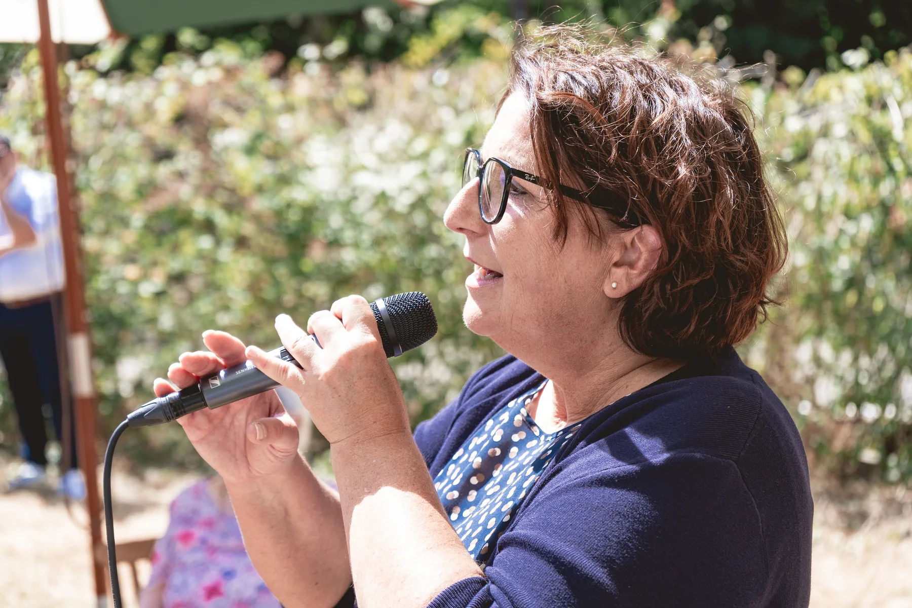 Petra Melzer spricht im Sonnenlicht ins Mikrofon, hinter ihr eine grüne Hecke